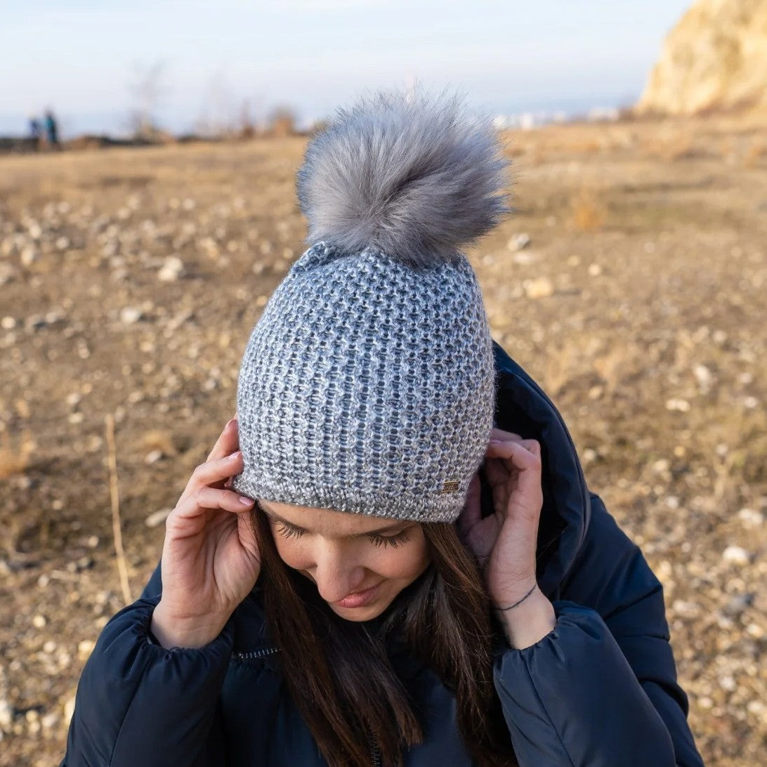A woman outdoors on a sunny winter day wearing the SABBOT Radka Bobble Hat in grey