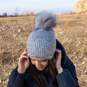 A woman outdoors on a sunny winter day wearing the SABBOT Radka Bobble Hat in grey