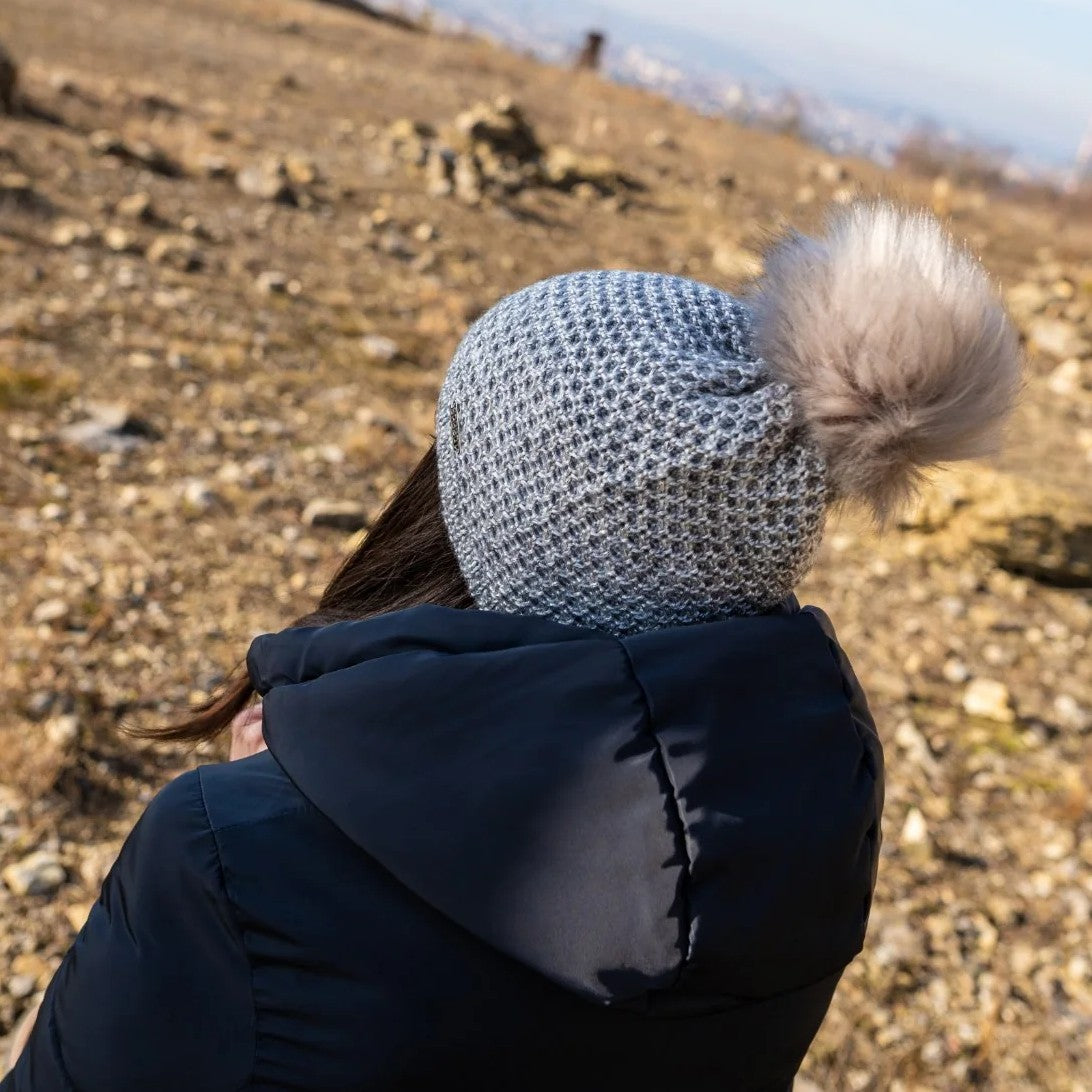 A woman sitting on a hillside, seen from behind, wearing the SABBOT Radka Bobble Hat in grey