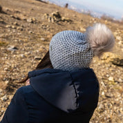 A woman sitting on a hillside, seen from behind, wearing the SABBOT Radka Bobble Hat in grey