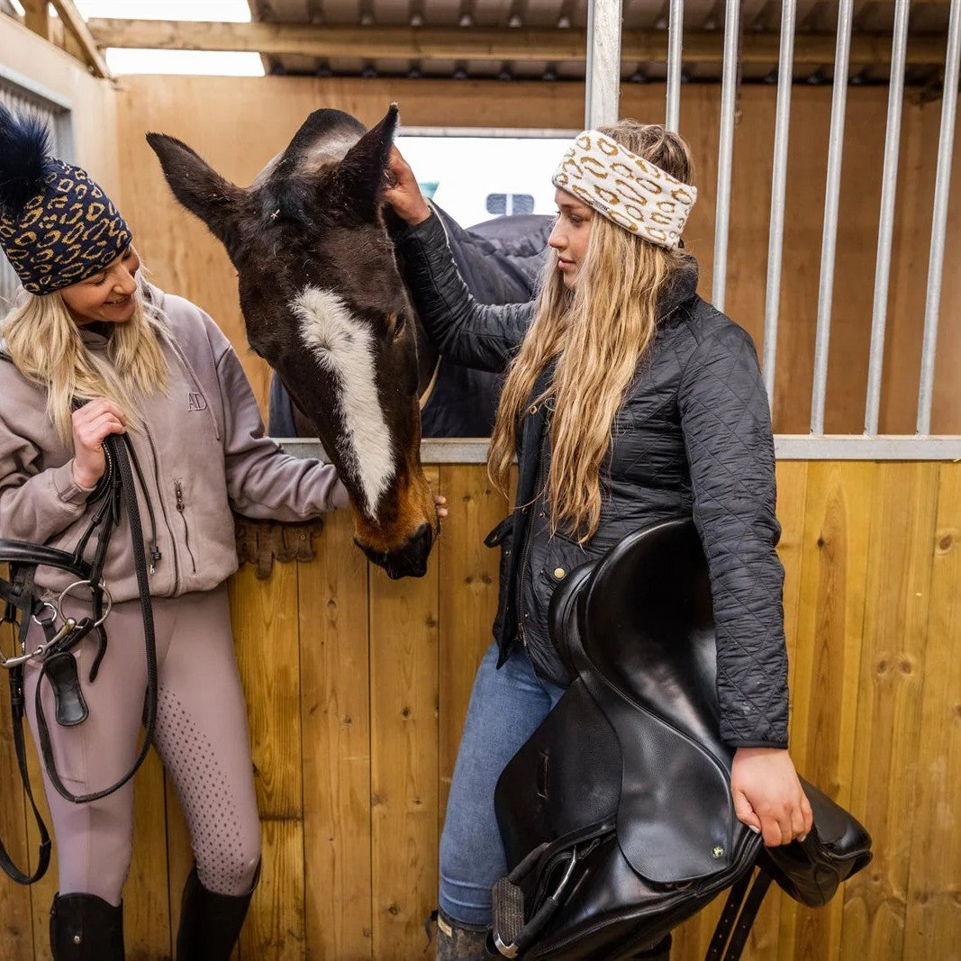 Friends with mugs in a tack room and stables wearing the Saskia Bobble Hat in Black/Gold and Saskia Headband in White/Gold