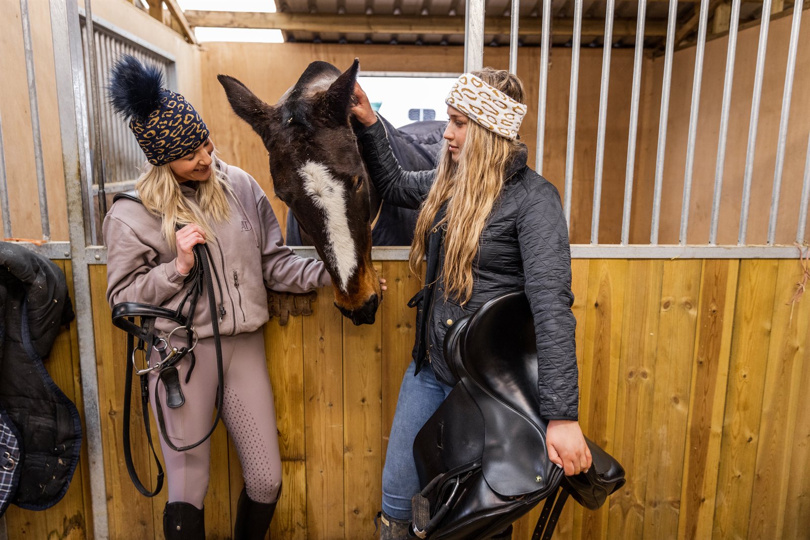 Friends with mugs in a tack room and stables wearing the Saskia Bobble Hat in Black/Gold and Saskia Headband in White/Gold