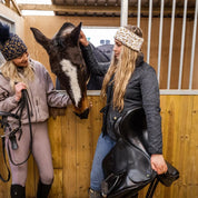 Friends with mugs in a tack room and stables wearing the Saskia Bobble Hat in Black/Gold and Saskia Headband in White/Gold