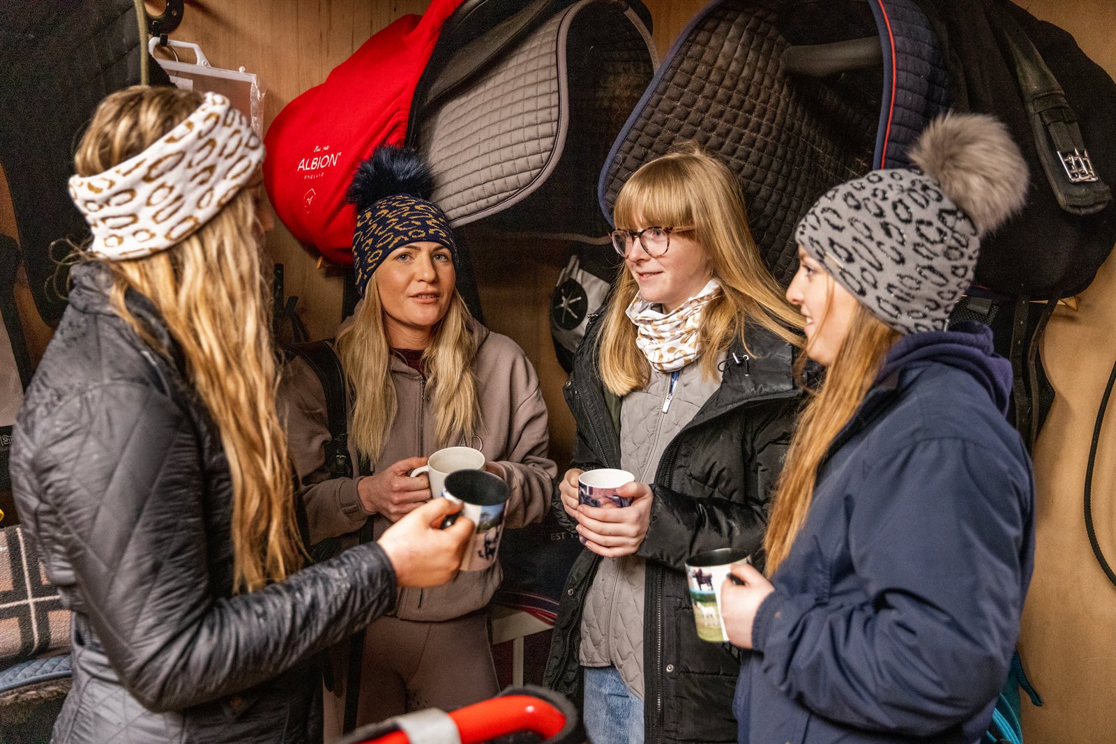 Friends with mugs in a tack room and stables wearing the Saskia Bobble Hat and Headband in leopard print