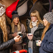 Friends with mugs in a tack room and stables wearing the Saskia Bobble Hat and Headband in leopard print