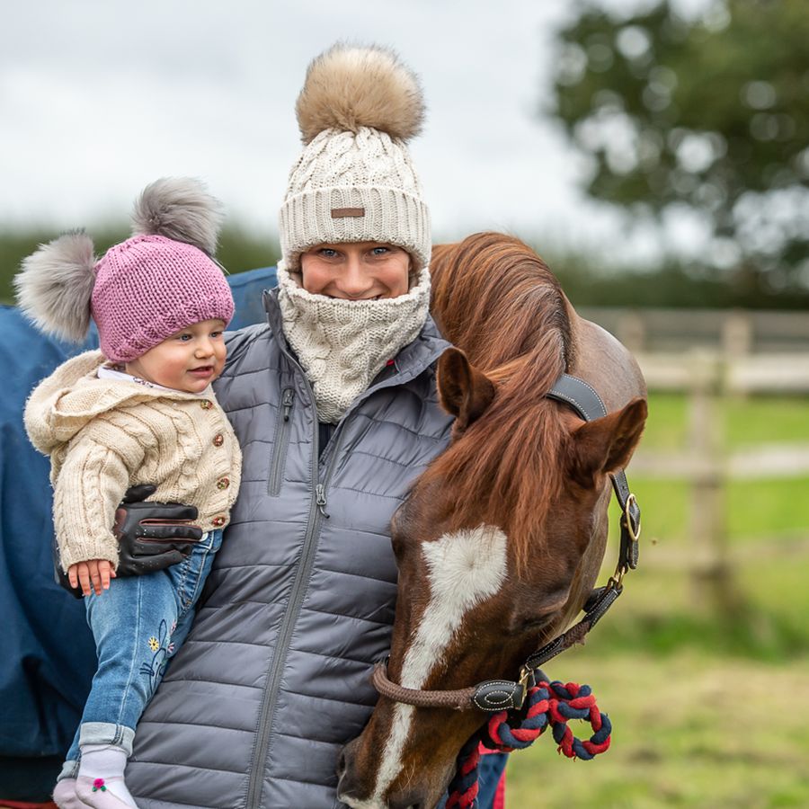 Woman at the stables in a cream cable knit SABBOT Linda bobble hat and matching snood, holding a toddler in a pink Frankie double pom-pom hat beside a horse