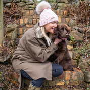 Woman in a pink SABBOT Petra Fold bobble hat with cream faux fur pom-pom hugging her spaniel by a stone wall