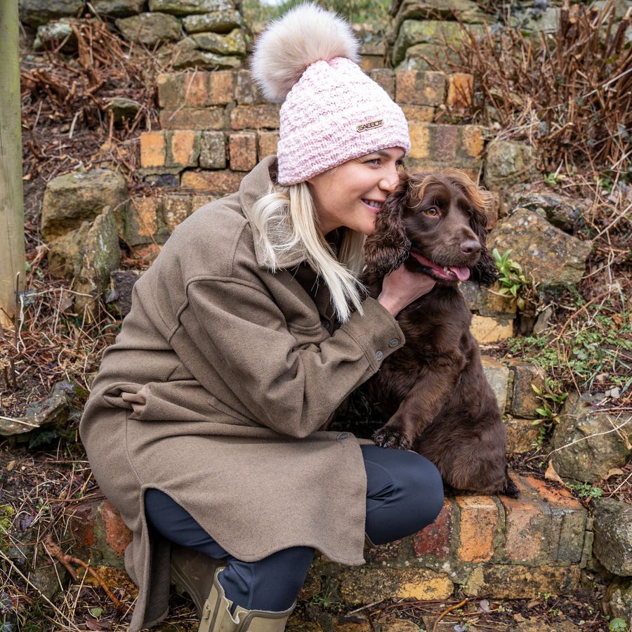 Woman in a pink SABBOT Petra Fold bobble hat with cream faux fur pom-pom hugging her spaniel by a stone wall