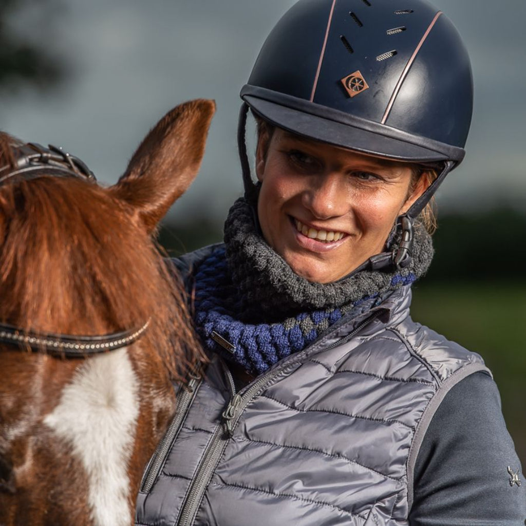 Smiling rider in a riding helmet wearing a SABBOT Chloe ombre knit snood next to her horse