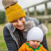Mum in a mustard chunky knit SABBOT Eva bobble hat with faux fur pom-pom cuddling a baby in a white Frankie double pom-pom hat